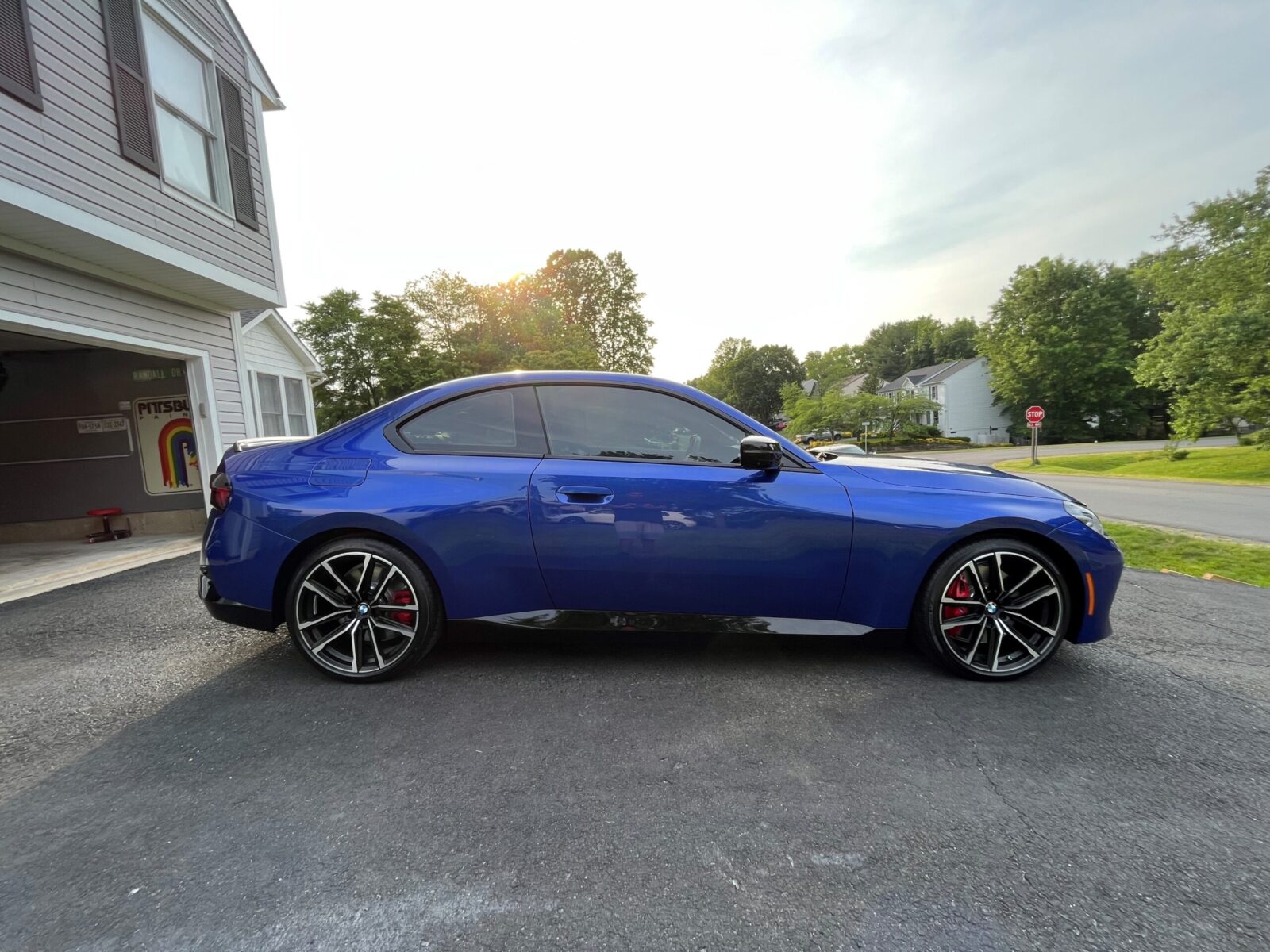 A blue sports car is parked on a driveway in a suburban neighborhood, taken in broad daylight.