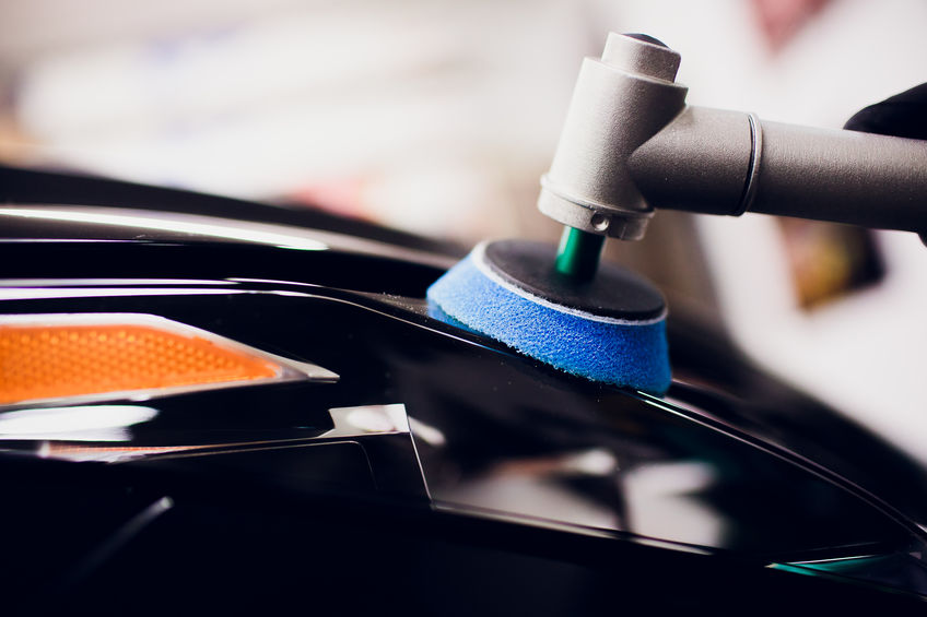 Close-up view of a person using a machine to polish a black car, with a blue foam pad making contact with the car's surface.