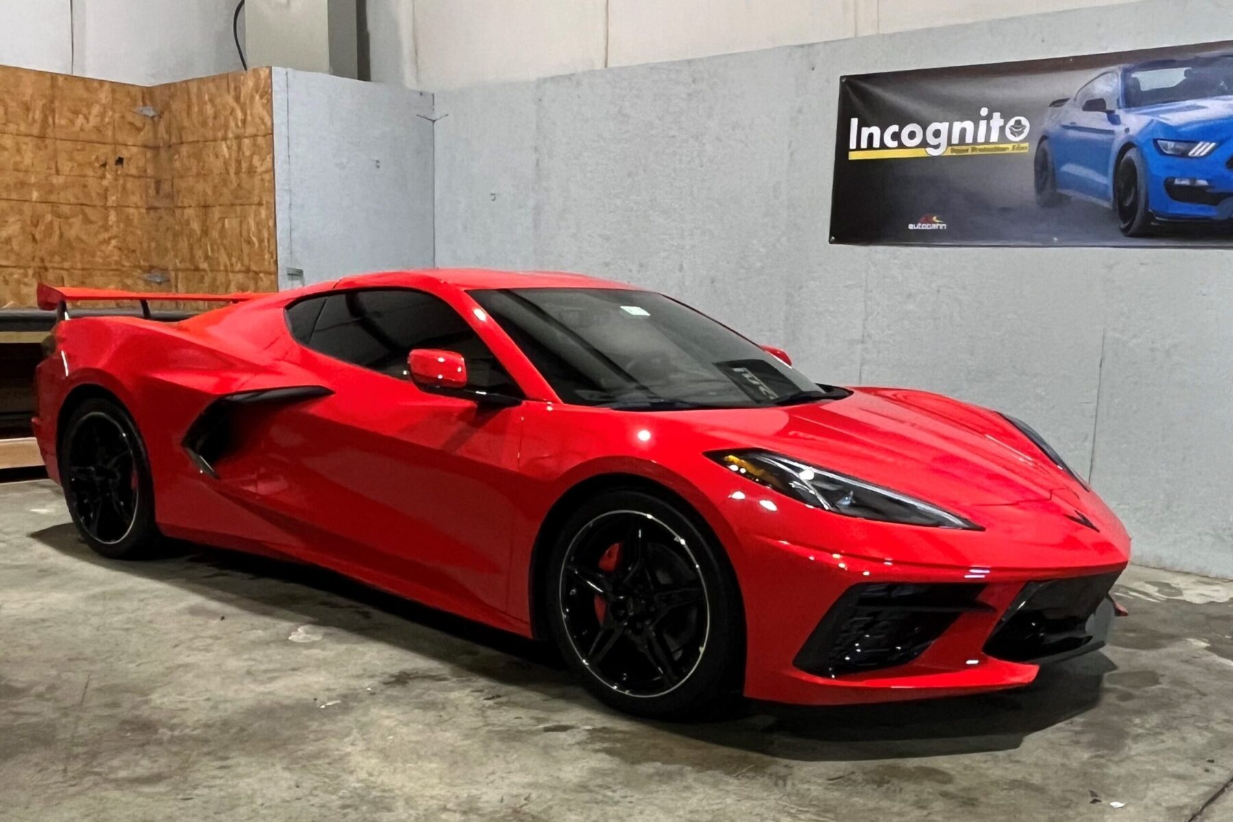 A red sports car is parked indoors next to a gray wall with a poster displaying a blue car and the word "Incognito.