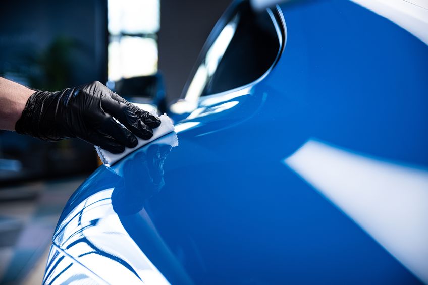 A gloved hand wiping and polishing the shiny blue surface of a car with a white cloth.