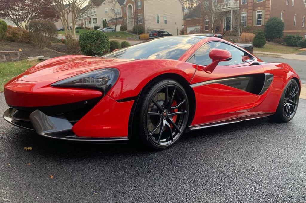 A sleek red sports car is parked on a residential street with houses and greenery in the background.