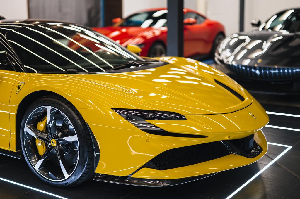 A close-up of a yellow sports car displayed in a showroom, with red and dark gray cars visible in the background.