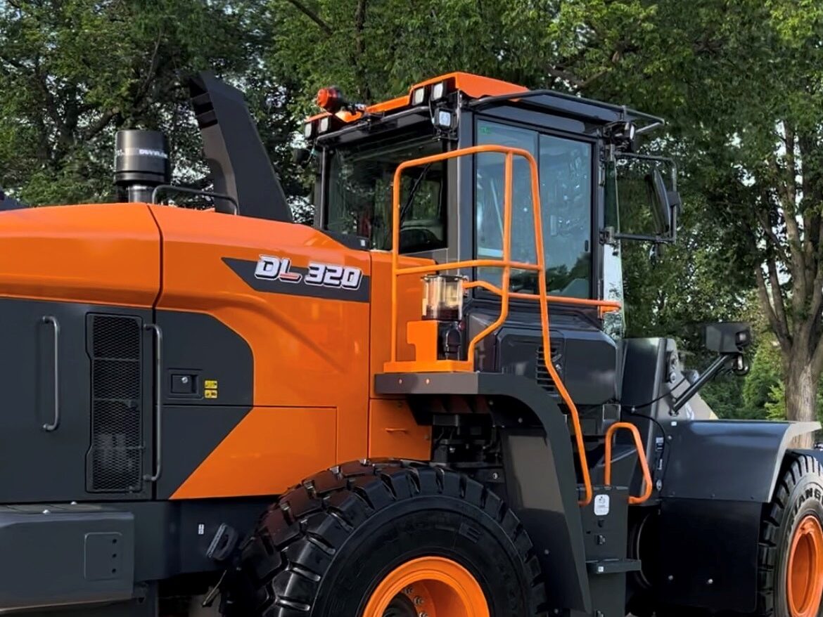 A large orange and black construction vehicle labeled "DL320" is parked outdoors with trees in the background.