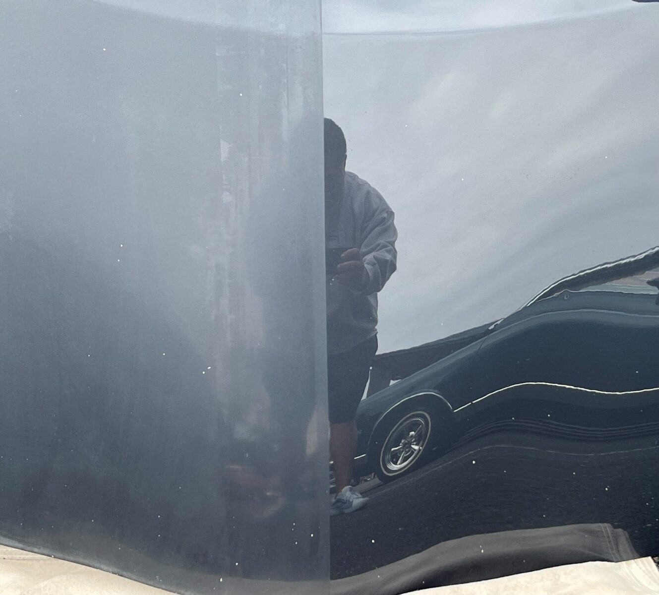 Reflection of a person in a gray jacket taking a photo in a shiny, slightly distorted surface of a car.