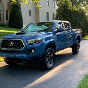 A blue pickup truck is parked on a driveway in front of a beige house surrounded by greenery.