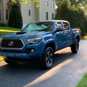 A blue pickup truck is parked on a driveway in front of a beige house surrounded by greenery.
