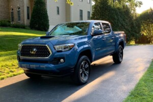 A blue pickup truck is parked on a driveway in front of a beige house surrounded by greenery.