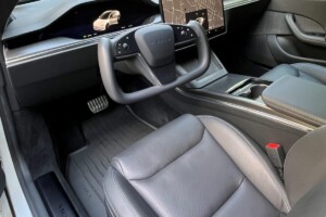 Interior of a Tesla vehicle showing the steering wheel, dashboard with digital displays, and front seats in a sleek, modern design.